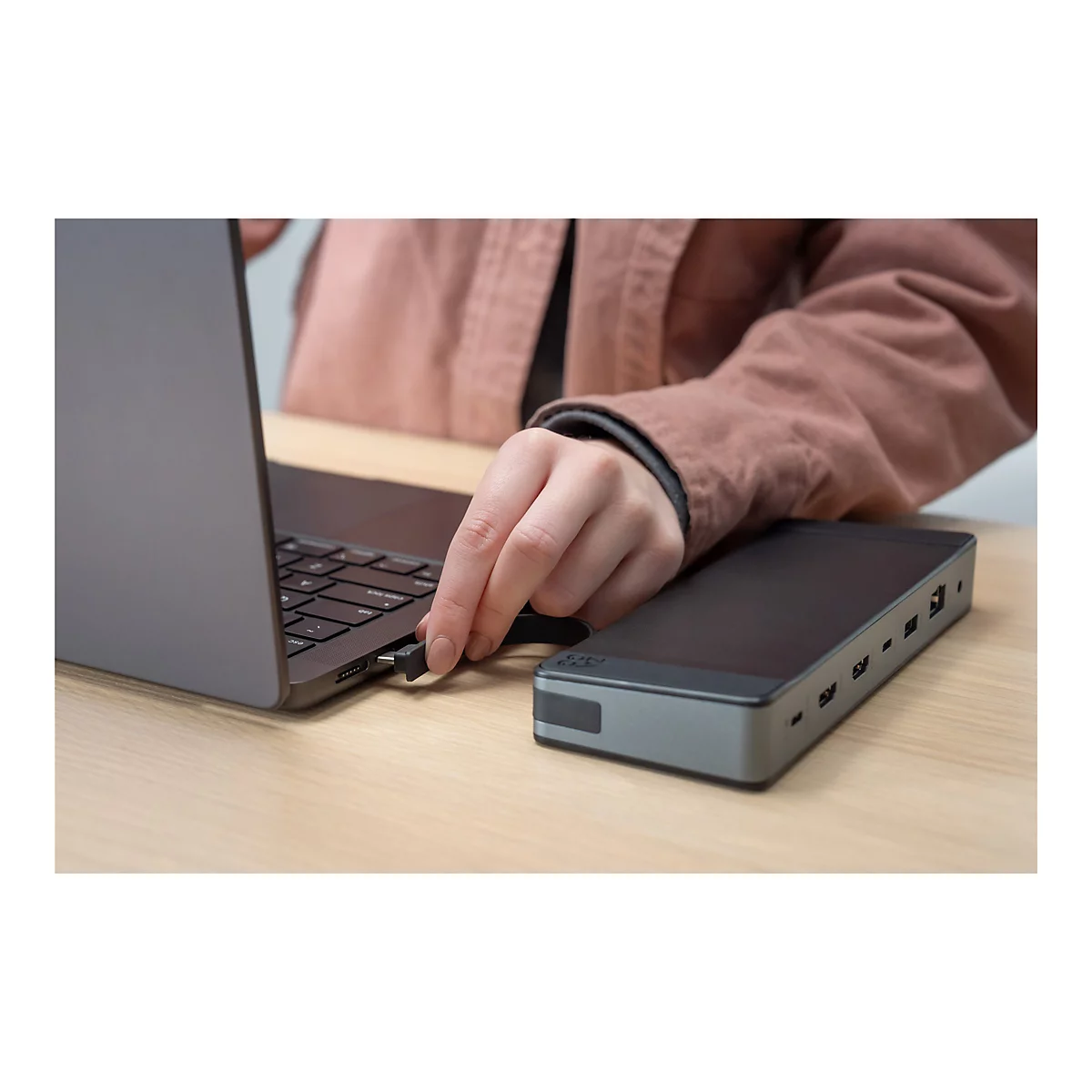Une personne connecte un connecteur USB-C à un ordinateur portable. Une station d'accueil noire se trouve à droite. Le fond est une table en bois.