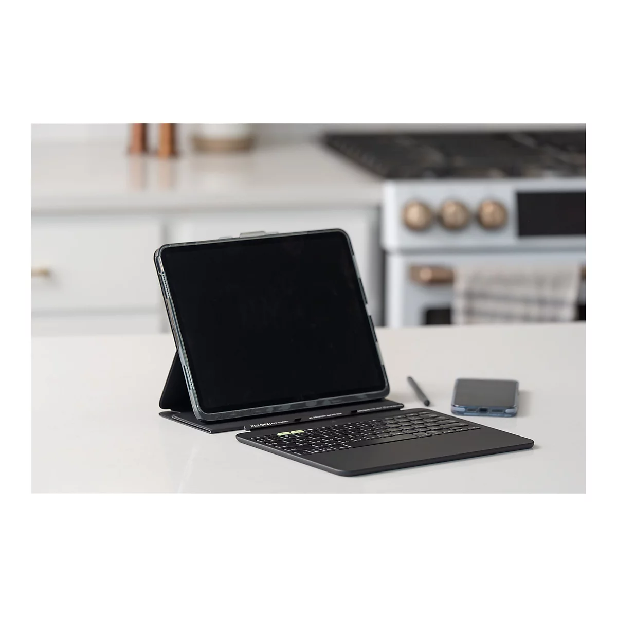 Ein Tablet mit Tastatur, Stift und Smartphone auf einer weißen Oberfläche. Im Hintergrund ist eine Küche unscharf zu sehen.