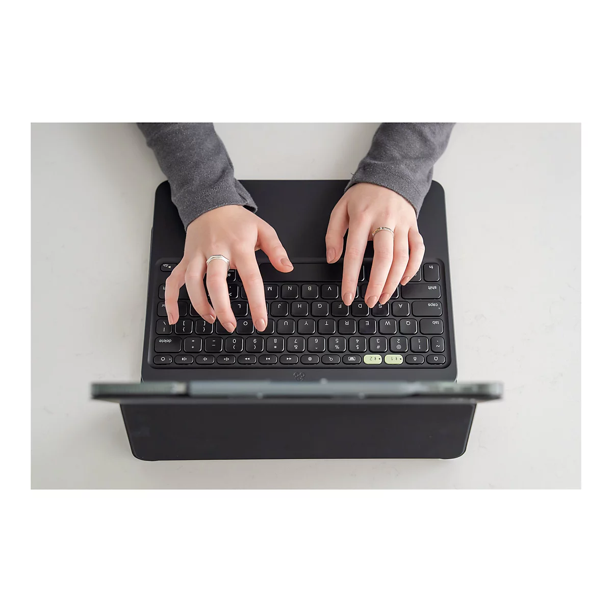 Nahaufnahme von Händen, die eine schwarze Tastatur bedienen. Die Person trägt einen grauen Pullover und Ringe an den Fingern. Die Tastatur ist mit einem Tablet verbunden und liegt auf einer weißen Oberfläche.