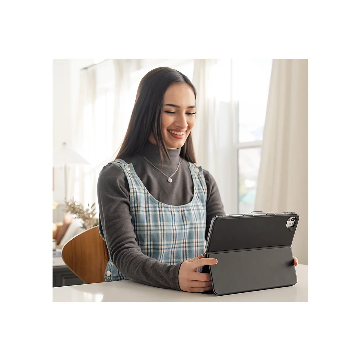 Lächelnde junge Frau, die auf einem Tablet sitzt. Sie trägt einen grauen Rollkragenpullover und eine blaue Latzhose. Auf dem Tisch befindet sich eine Vase mit dekorativen Ästen.