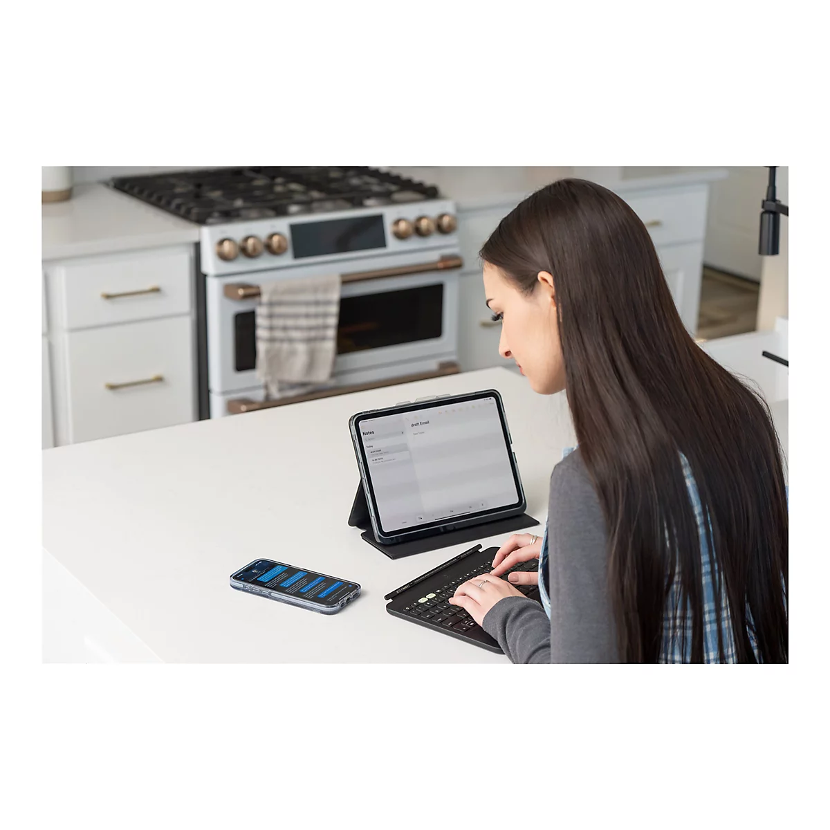 Eine Frau sitzt an einer Küchenarbeitsplatte und schreibt auf einer Tablet-Tastatur. Auf dem Tablet ist eine E-Mail zu sehen; daneben liegt ein Smartphone.