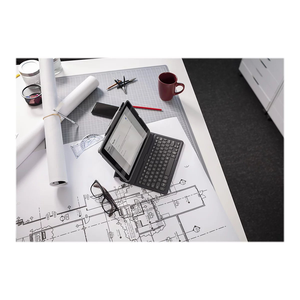 Draufsicht eines Schreibtisches mit einem Tablet mit Tastatur, Plänen, Stiften und einer Tasse.