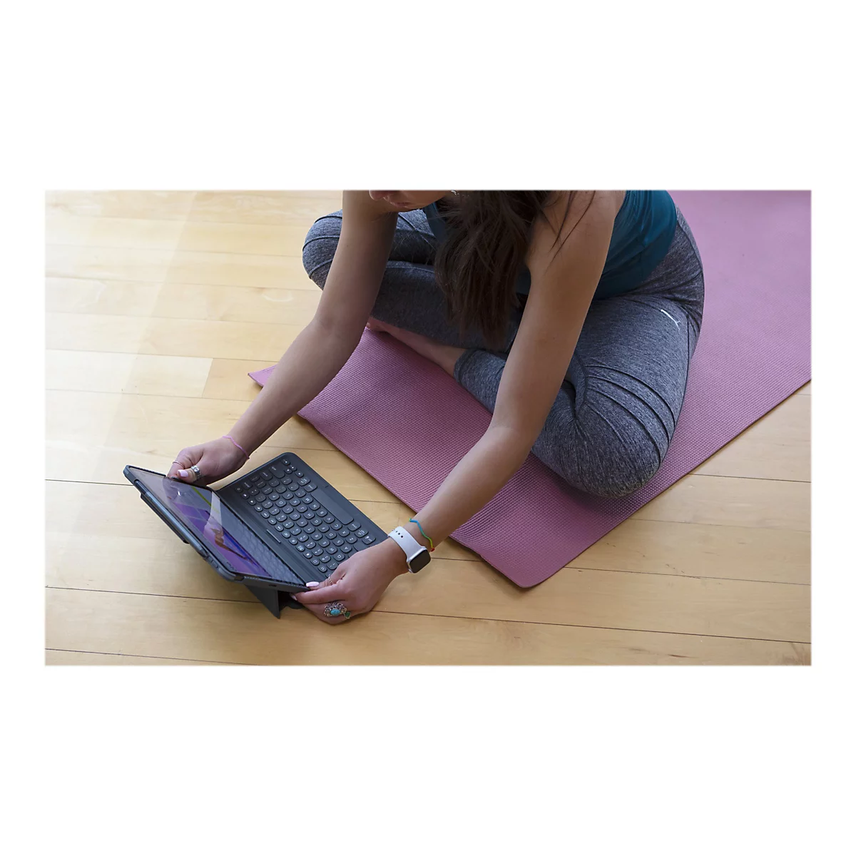 Eine Person in Yoga-Kleidung sitzt mit verschränkten Beinen auf einer Yogamatte und bedient ein Tablet mit Tastatur.