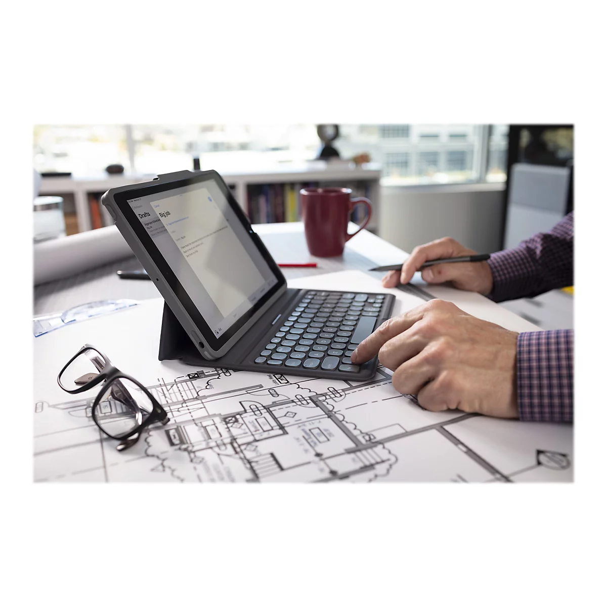 Ein Mann, der an einem Tisch mit einem Tablet-PC und Tastatur arbeitet, mit Bauplänen, einer Kaffeetasse und einer Brille.
