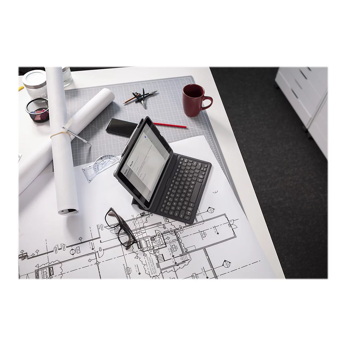 Ein Tablet mit Tastatur liegt auf einem Schreibtisch mit Bauplänen, daneben Brille und roter Becher.