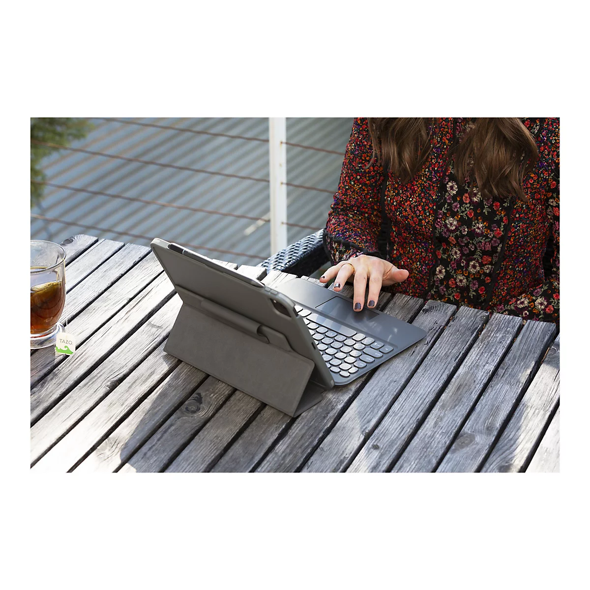 Une femme est assise à une table en bois et utilise une tablette grise avec un clavier. Sur la table se trouvent une tasse de thé et un sachet de thé avec l'inscription TAZO.