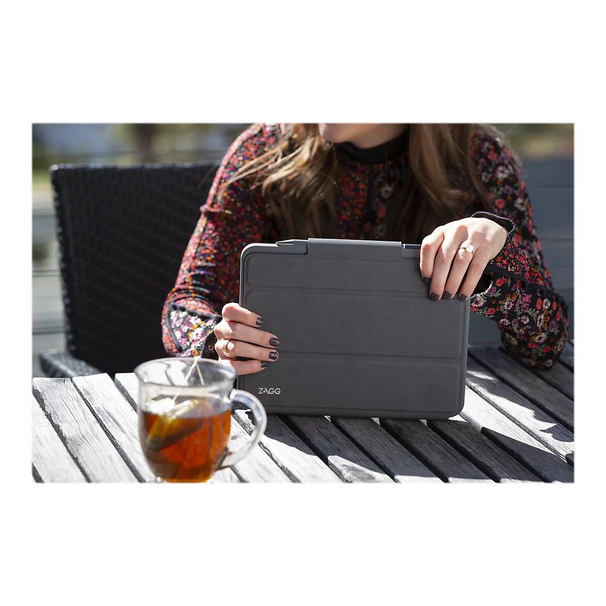 Une femme tient un étui de tablette gris sur une table en bois, à côté d'une tasse de thé.