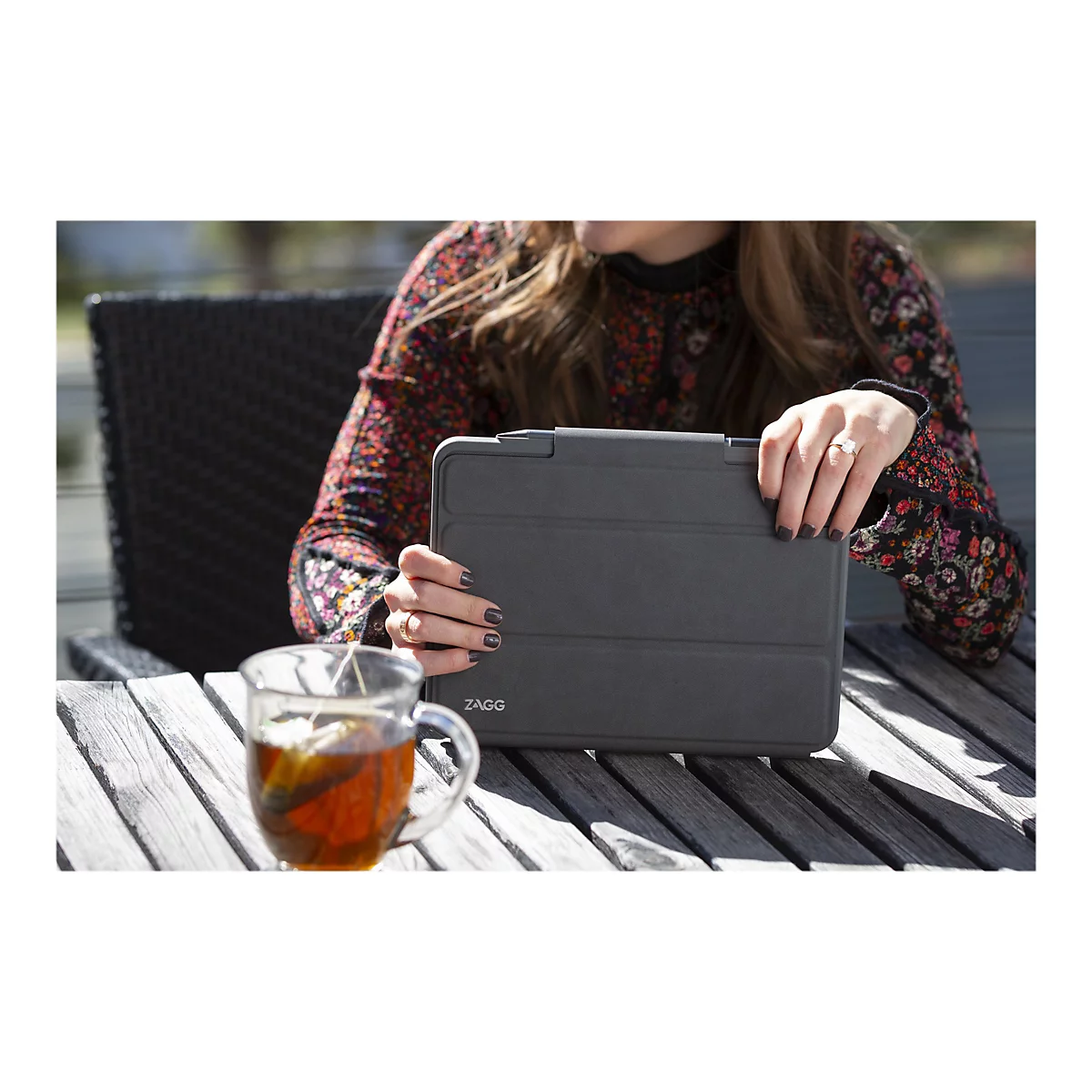 Une personne tenant un étui de tablette gris, à l'extérieur, à côté d'une tasse de thé.