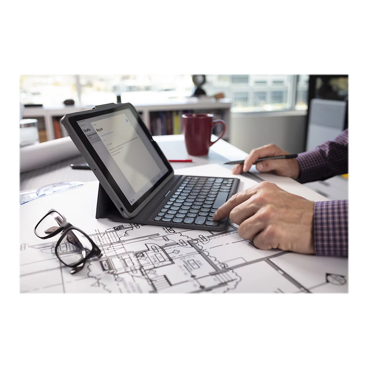 Person mit Tablet und Tastatur arbeitet an Bauplänen; Brille liegt daneben.