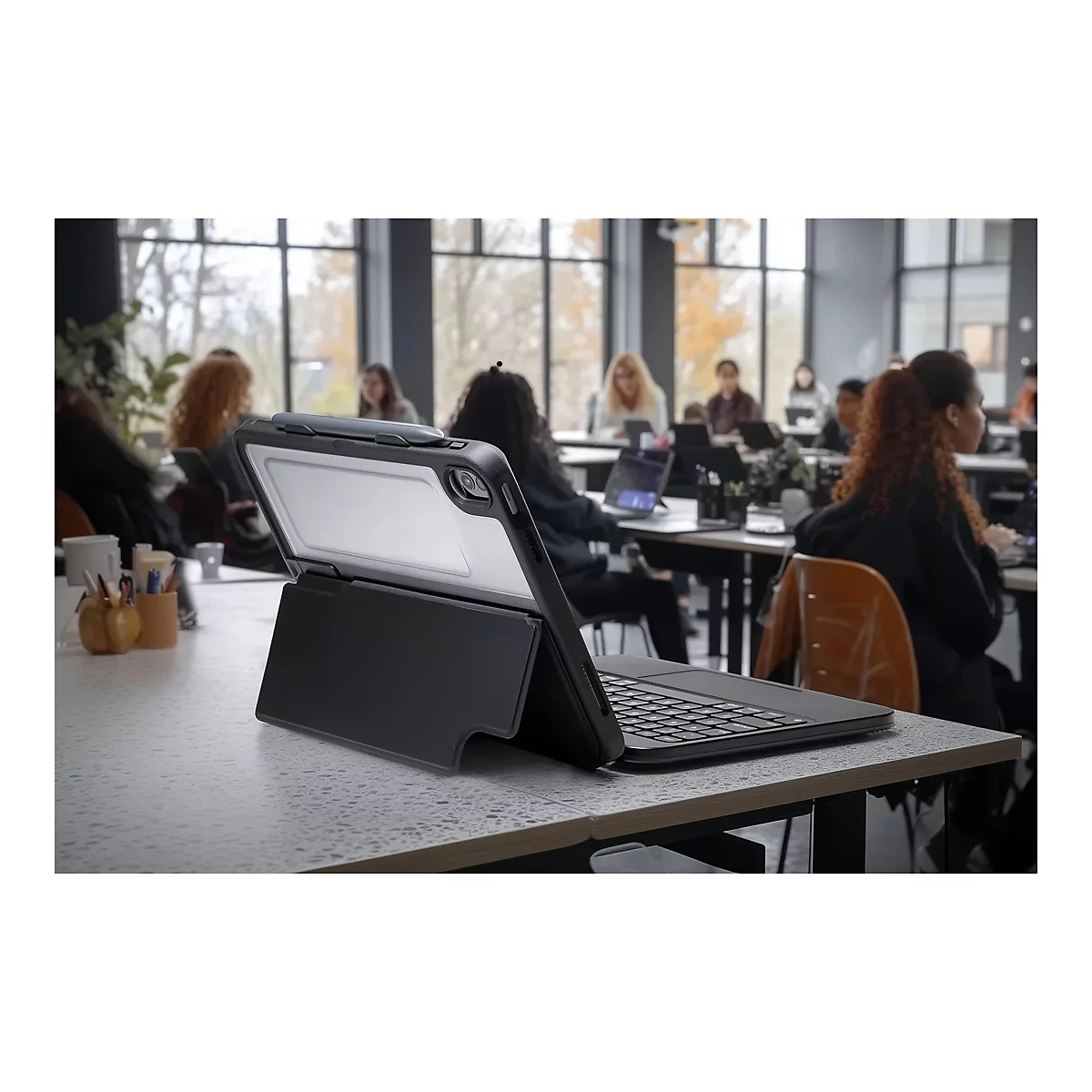 Schwarzes Tablet mit Tastatur auf einem Tisch in einem Büro. Im Hintergrund arbeiten mehrere Personen.