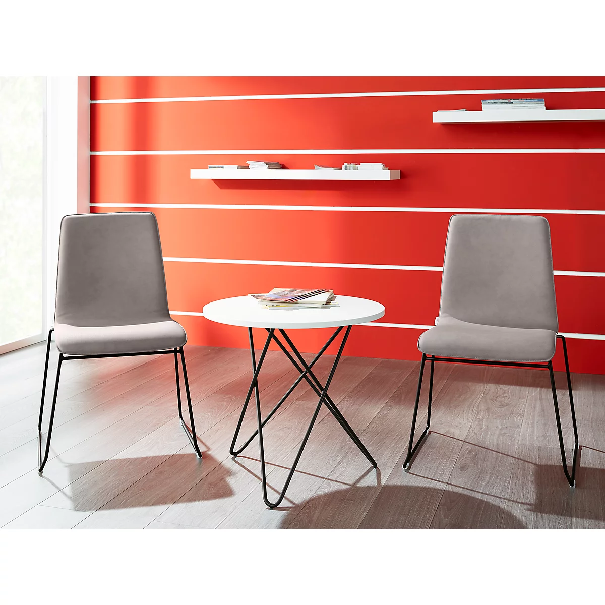 Two gray chairs with black legs surround a small white table in front of a red wall. Bookshelf and decoration.