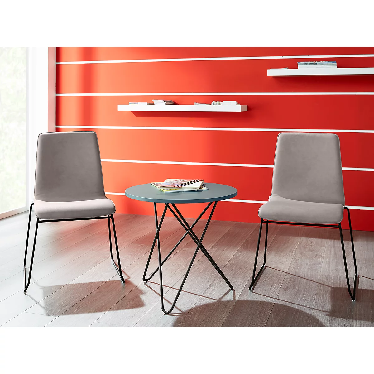 Two grey chairs in front of a red wall with white horizontal lines. In between, a small round table with magazines.