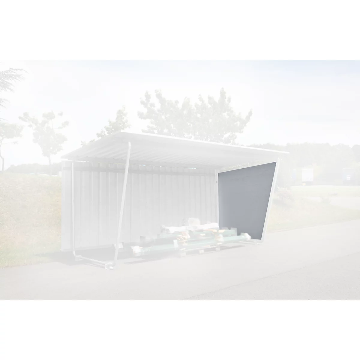 Een grijze schuur met metalen wanden en dak, gevuld met bundels hout, staat op asfalt. Bomen op de achtergrond.