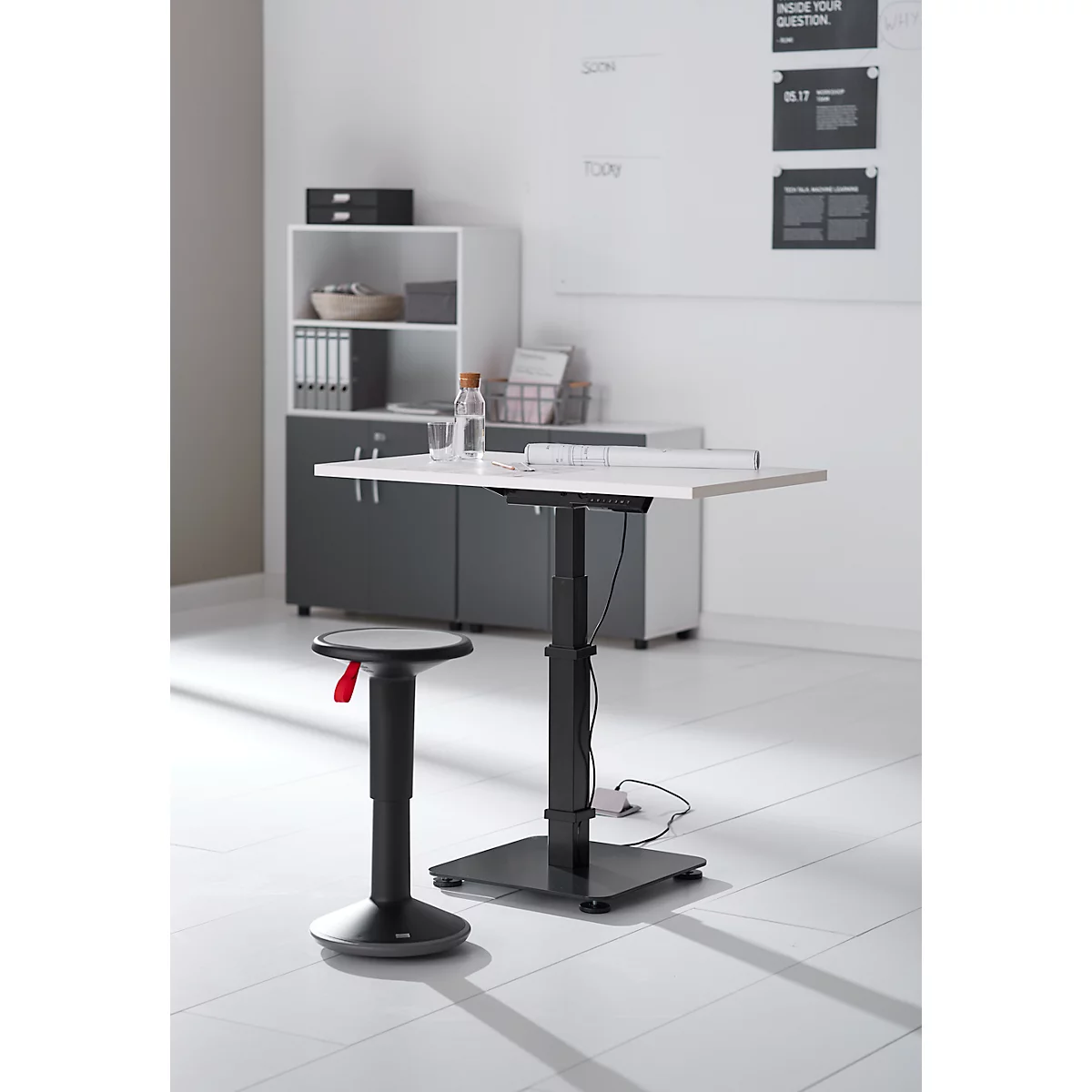 Een in hoogte verstelbaar bureau met een kruk in een kantoor. Er liggen documenten en een waterfles op de tafel.