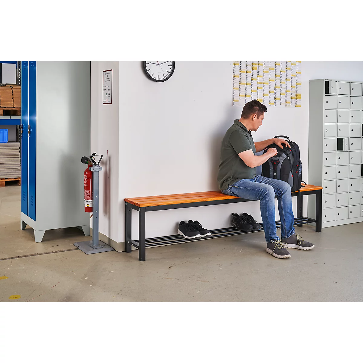 Een man zit op een bank en pakt zijn rugzak uit. Schoenen liggen op de onderste plank van de bank.