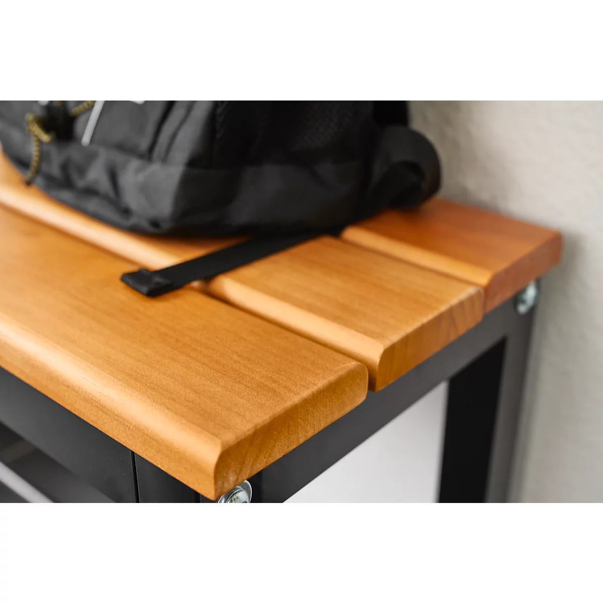 Detail van een houten bank met een zwarte rugzak erop. De bank heeft een bruin houten bovenblad en een zwart metalen frame.