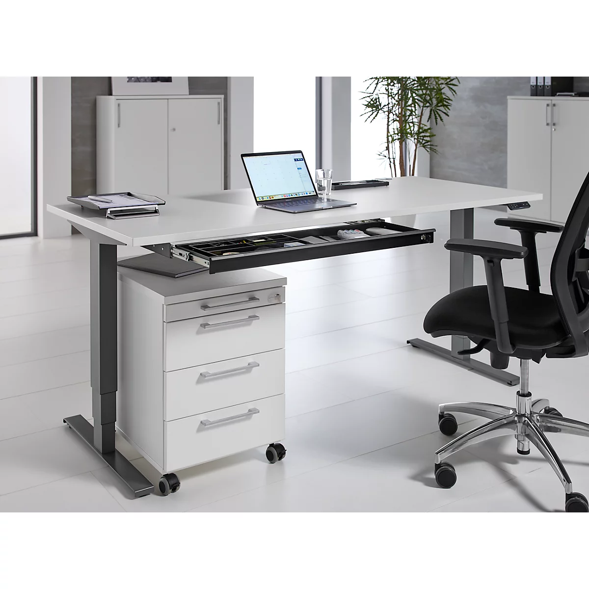 Weißer Schreibtisch mit Laptop, Stifthalter und Schublade, daneben ein schwarzer Bürostuhl.