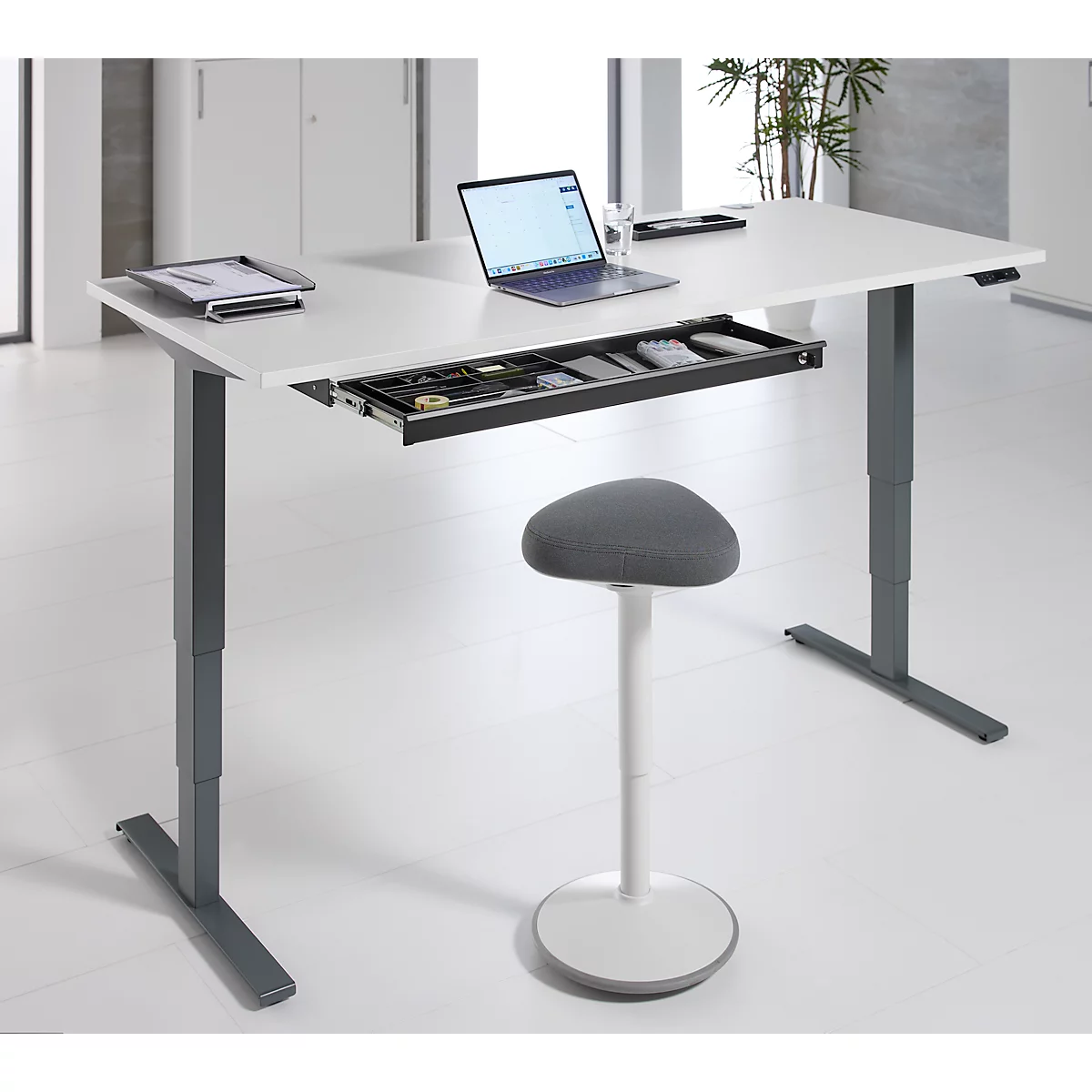 Ein höhenverstellbarer Schreibtisch mit Laptop, Schublade und Hocker. Auf dem Tisch liegen weitere Utensilien.